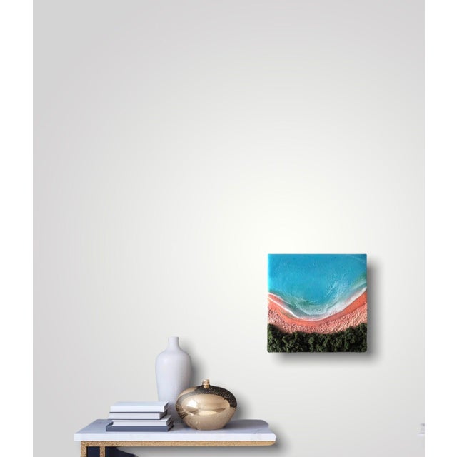 This mixed media piece captures a serene beach scene, where vibrant turquoise waters meet a soft, coral-hued shore. The...