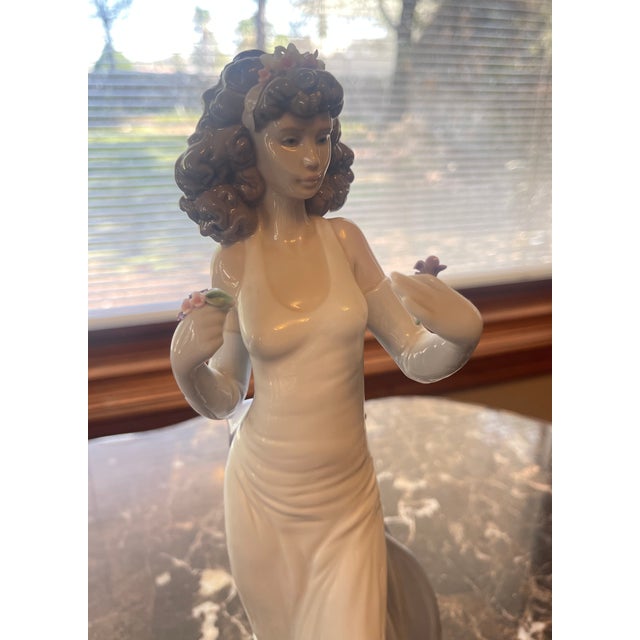Lladro female figurine with curly brown hair, wearing a floral headband and holding flowers. She is wearing a slinky white...