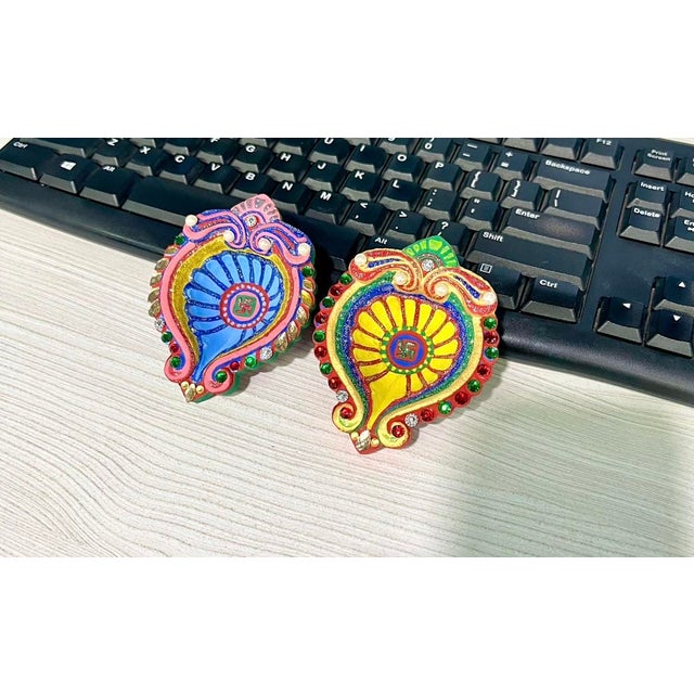 A colorful diya with a traditional swastika symbol sits beside a laptop, phone, and playing card—capturing the blend of...