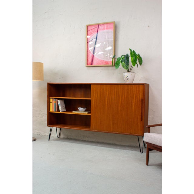 A beautiful teak highboard from the 1960s, immediately striking thanks to its slender, black hairpin legs. The warm brown...