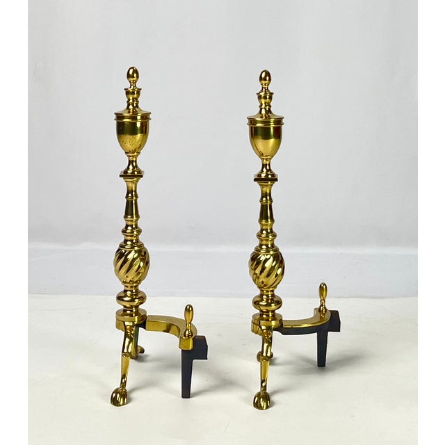 A striking pair of polished brass fireplace andirons featuring traditional urn finials above a bold spiral-twist (flame-...