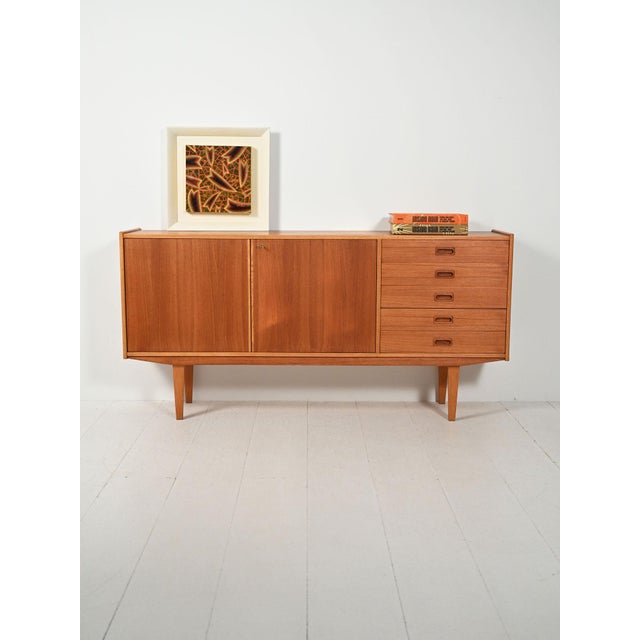 A 1960s teak sideboard with two doors and drawers with recessed handles, raised profiles, and sleek legs. A 1960s...