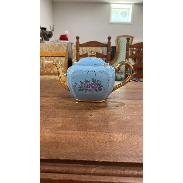 An charming vintage light blue porcelain teapot featuring a lobed body adorned with hand-painted pink rose bouquets and...