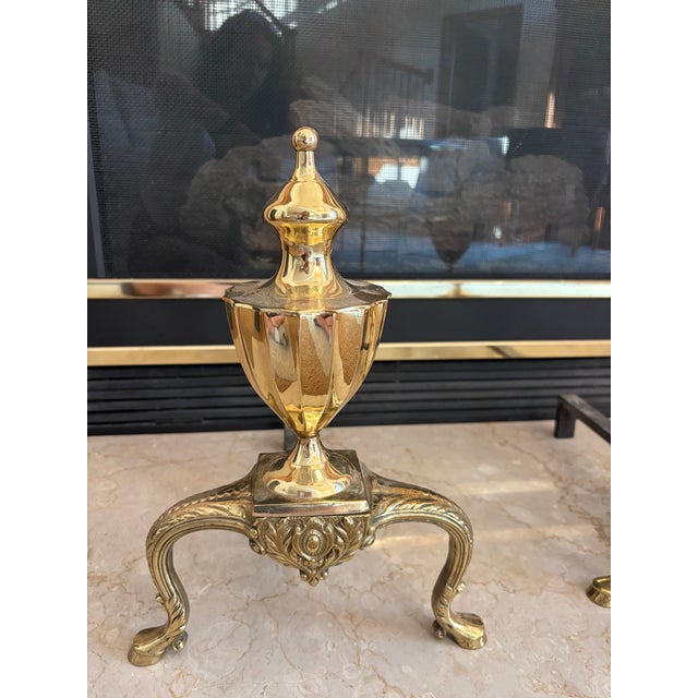 A decorative pair of vintage 1990s brass fireplace andirons in the Colonial Revival style. Each features a polished urn-...