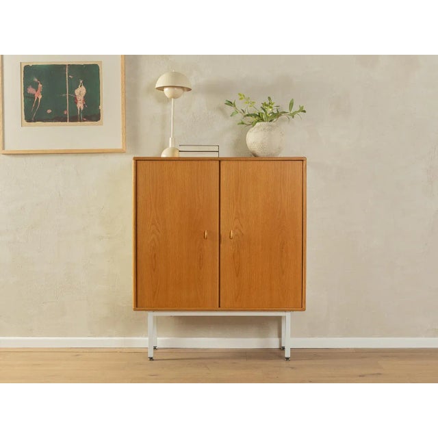 Body in oak veneer with two doors, a shelf and new square steel feet in white. The sides were coated with Formica in cream...