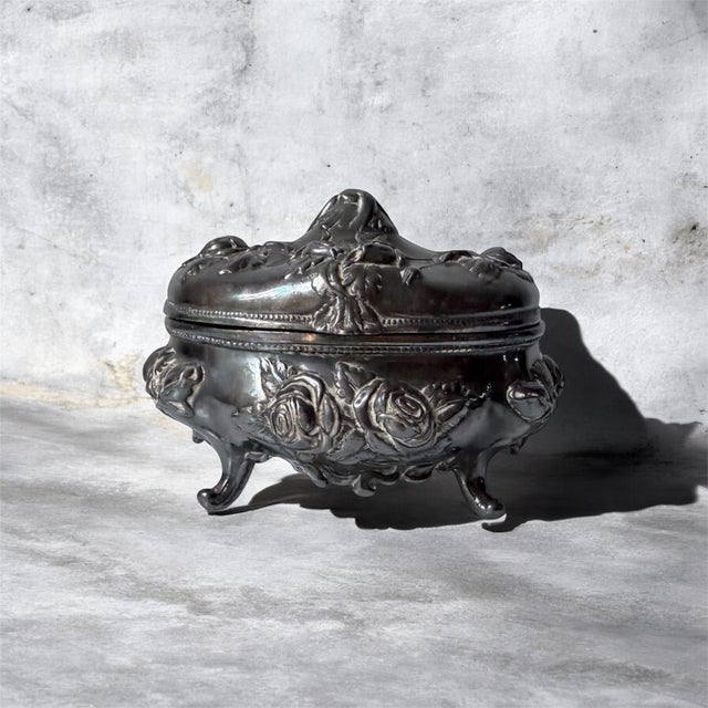 This antique silver-plated jewelry casket features a hinged lid and an ornate floral design, typically associated with the...