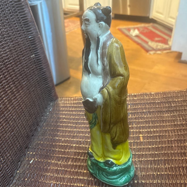 This antique Chinese porcelain figurine likely depicts a Taoist immortal or deity, such as Zhongli Quan holding a fan. It...