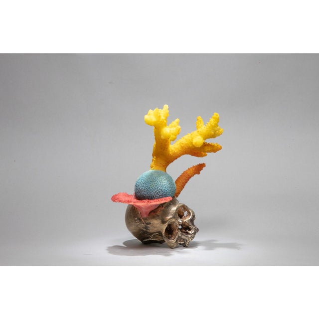 This sculpture features a striking juxtaposition of a skull rendered in bronze and resin, adorned with vibrant coral-like...