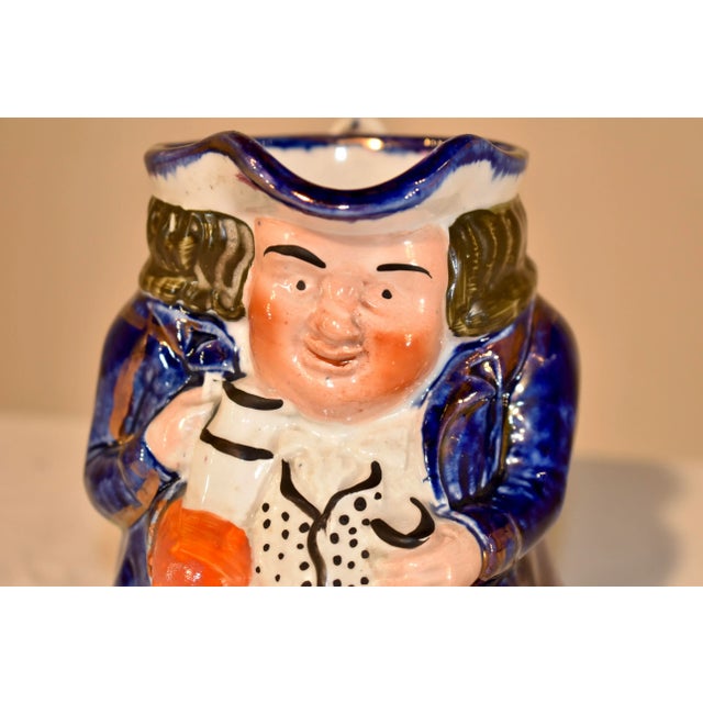19th century ceramic Toby jug from the Staffordshire region of England. He has a blue jacket and a warm expression on his...