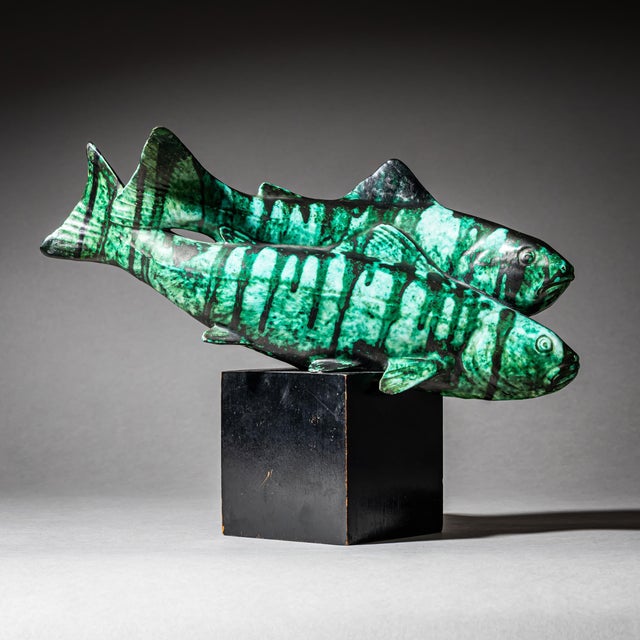 Ceramic sculpture depicting two trout with a green-black enameled surface. The group is mounted on a black cubic base....