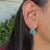 This elegant earring features a pair of 5.32 carat Sleeping Beauty Turquoise gemstones with a halo of natural earth mined...