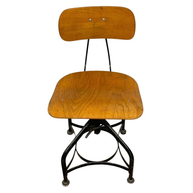 Vintage industrial drafting chair featuring a molded wood seat and backrest mounted on a black metal frame. The chair...