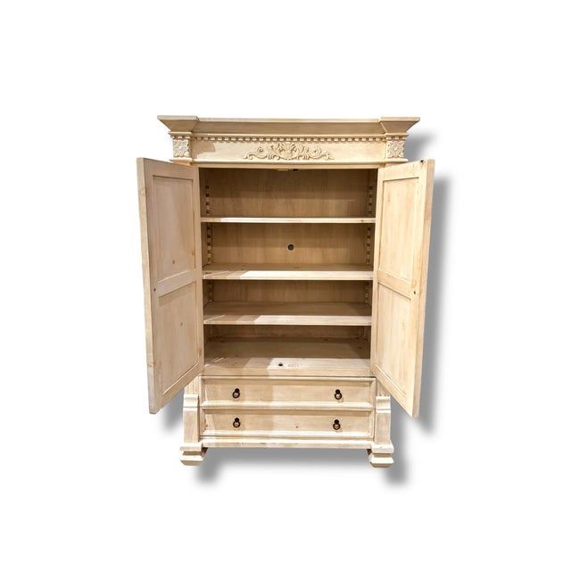 A beautifully detailed pre-loved armoire featuring a light natural wood finish and elegant carved accents along the crown...