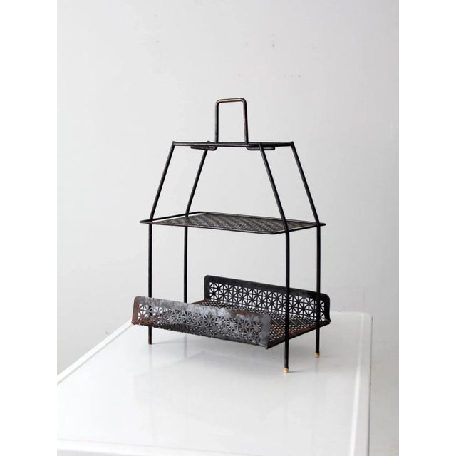 A mid-20th-century metal stand in a sleek black finish, featuring two shelves with an expressive patterned cutout design....