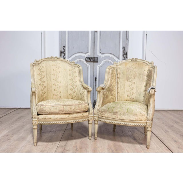 A pair of French Louis XVI style painted bergères à oreilles from circa 1900 with carved acanthus leaves and fluted legs....