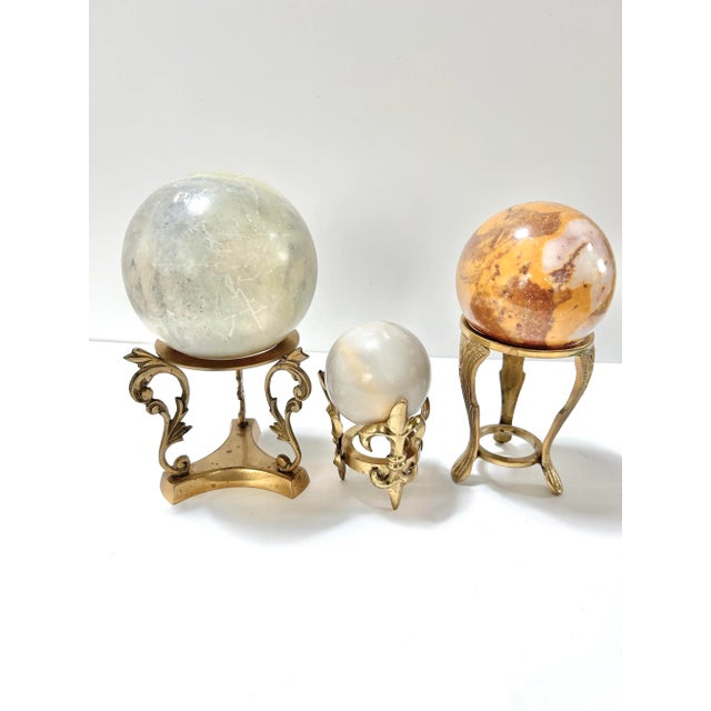 A sculptural set of three decorative stone spheres displayed on ornate brass pedestals. Each sphere features natural...