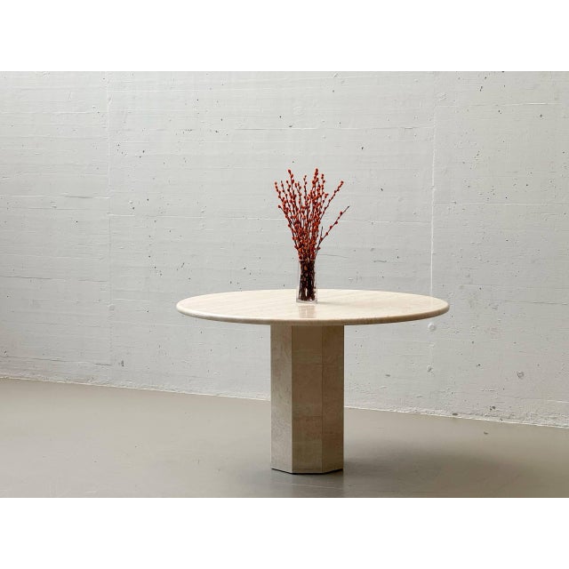 Sculptural dining table made of solid travertine with a round tabletop and a central, polygonal pedestal base. The...