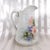 This is a white milk jug or pitcher with a multicolor floral design that has a floral design that goes around three...