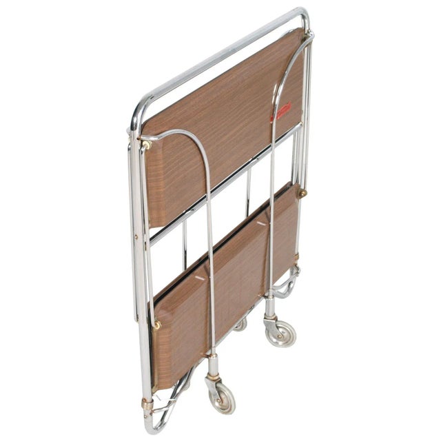 This foldable bar or serving cart was produced in Germany in the 1970s. It has a chrome frame with Formica trays, in teak...