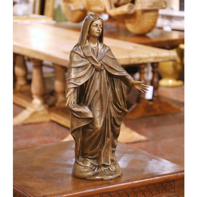 This refined antique bronze sculpture depicts the Virgin Mary in a serene, open-armed pose, symbolizing grace, compassion,...