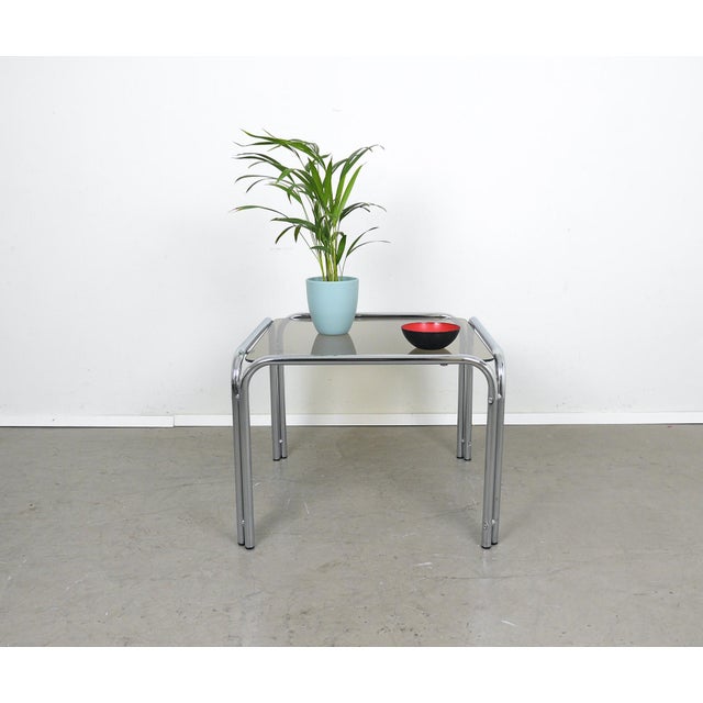 Side table from the 1970s. A square, lightly tinted glass top rests on an arched frame made of chrome-plated steel tubing.