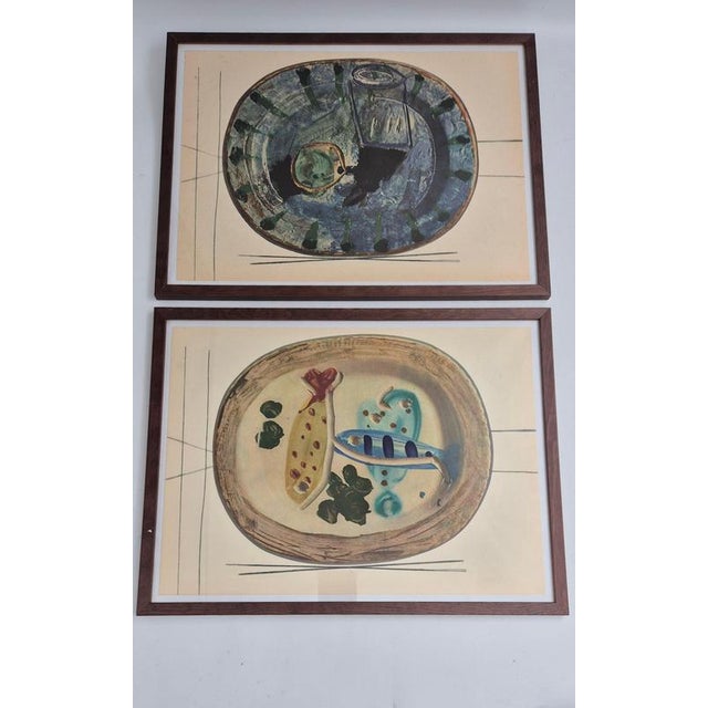 Two framed shiny polychrome prints of Picasso Vallauris ceramic plates depicting food. The color print is attached to a...