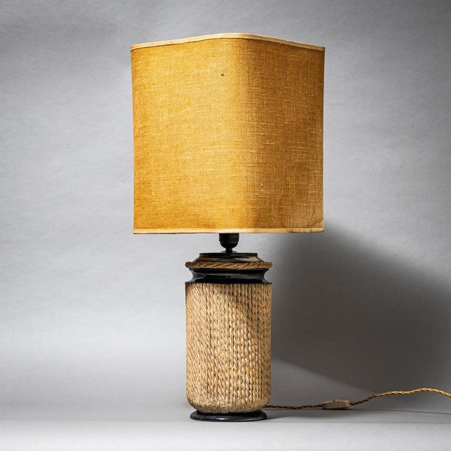 Table lamp featuring a black cylindrical ceramic base decorated with beige rope wrapping. The shade is made of beige woven...
