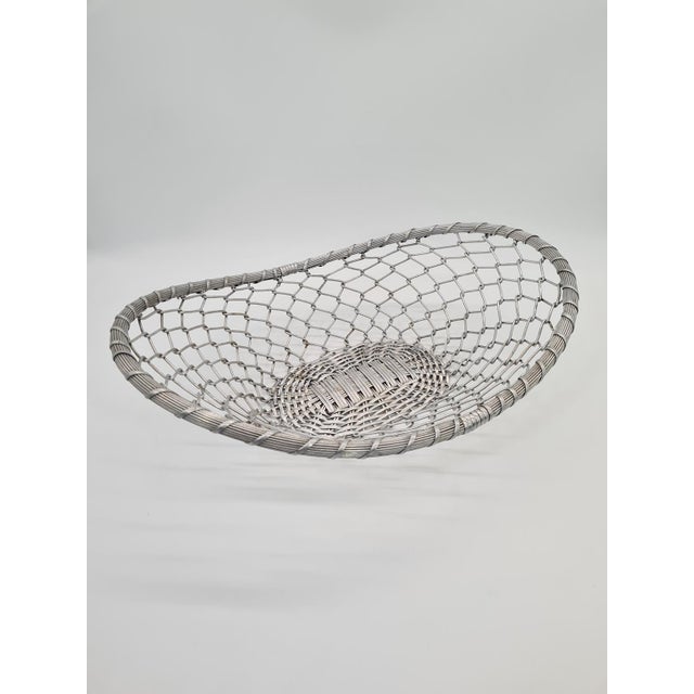 A decorative fruit basket made of bent aluminum, with a light and openwork structure resembling a braid. The form is based...
