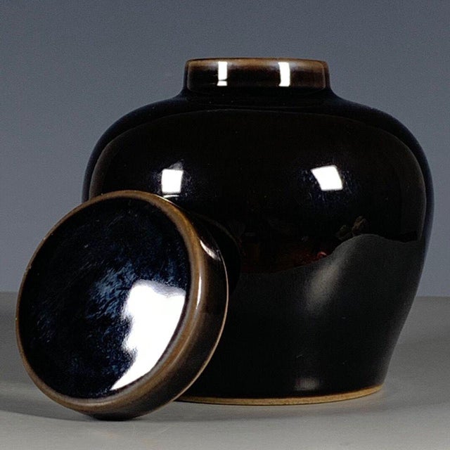 A sleek, glossy black glazed covered jar with a softly rounded belly and domed lid. The deep, mirror-like black glaze is...