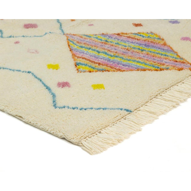 A vivid striped diamond glows at the top of this ivory rug like a woven sunburst, anchoring a playful scatter of tiny...
