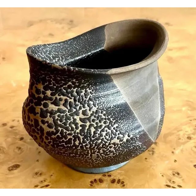 This ceramic vessel features a striking contrast of textures, with a rough, volcanic-like surface blending into smooth,...
