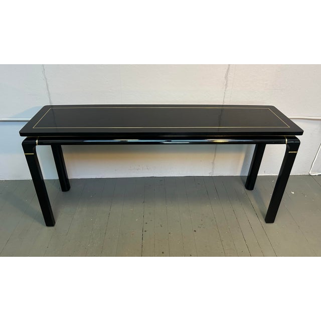 This vintage console table by John Stuart features a stylish Mid-Century design with dark burl lacquered finish and brass...