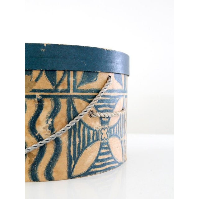 Vintage mid-century hat box featuring a round form, blue and tan graphic paper design, matching lid, and braided cord...