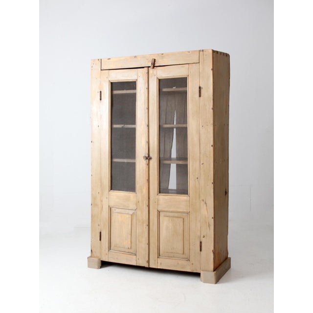 This antique 19th-century pine farmhouse cupboard features double doors with mesh wire windows that offer a glimpse...