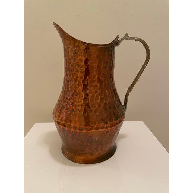 A sculptural vintage hammered copper pitcher featuring a warm, richly aged surface and cast brass handle. The vessel has a...