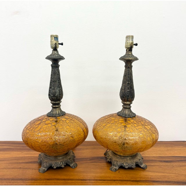 A striking pair of vintage table lamps featuring oversized amber crackle glass bodies complemented by ornate cast metal...