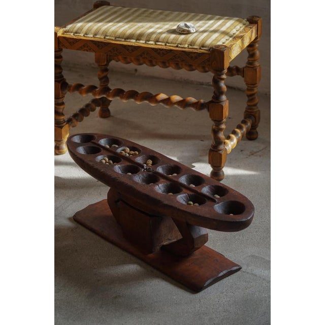 A hand-carved wooden mancala game board showcasing a sculptural and well-balanced form. The top features a series of...