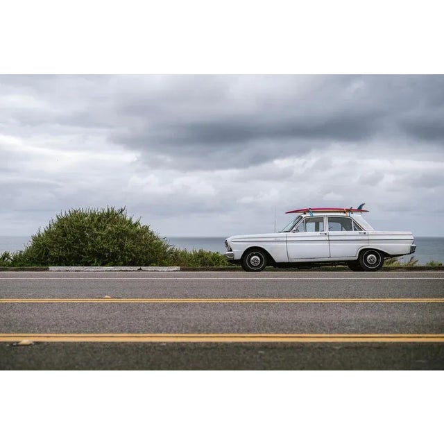 A 1960s Ford Falcon parked on Pacific Coast Highway 101 with a pink surfboard on top is waiting for sun on a grey...