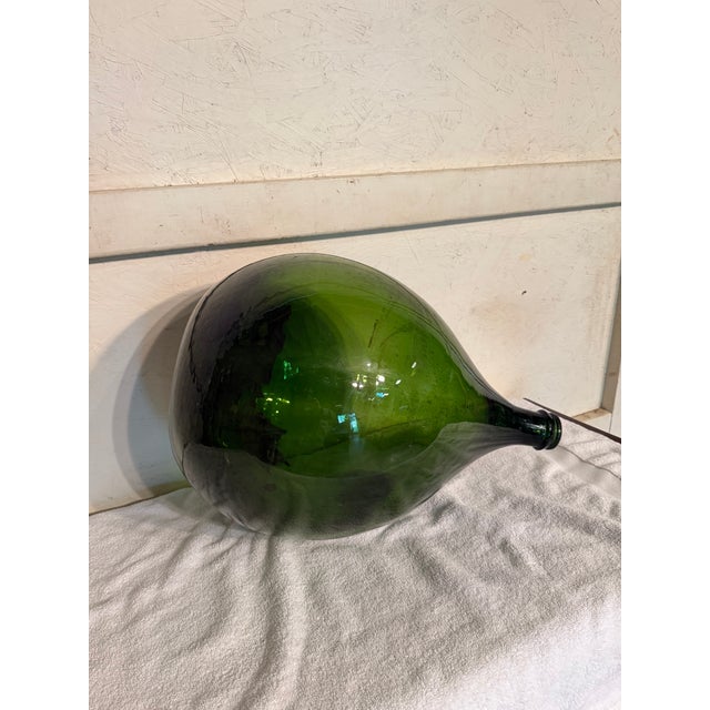 Vintage large green glass demijohn bottle with a rounded body and tapering neck. The bottle features remnants of an old...