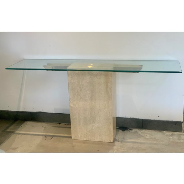 A vintage 1970s narrow console table consisting of a travertine rectangular base and a beveled glass top. The top is...