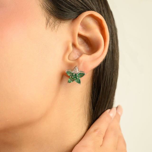 Elegant silver stud earrings showcasing vivid green emeralds arranged in a refined star-shaped floral cluster, accented...