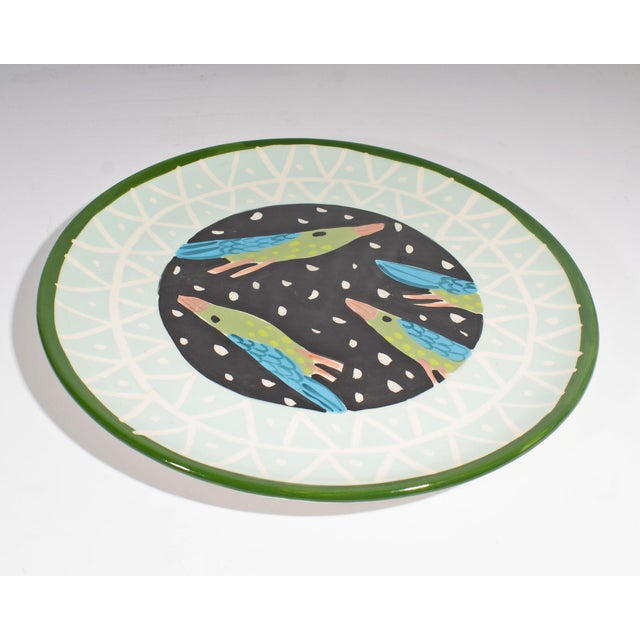A ceramic bird plate designed by Droll Designs. The center of the plate has four green and blue birds with orange beaks...