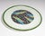 A ceramic bird plate designed by Droll Designs. The center of the plate has four green and blue birds with orange beaks...