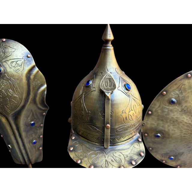 A three-piece brass armor display featuring a pointed helmet, round shield, and matching forearm guard mounted on a dark...