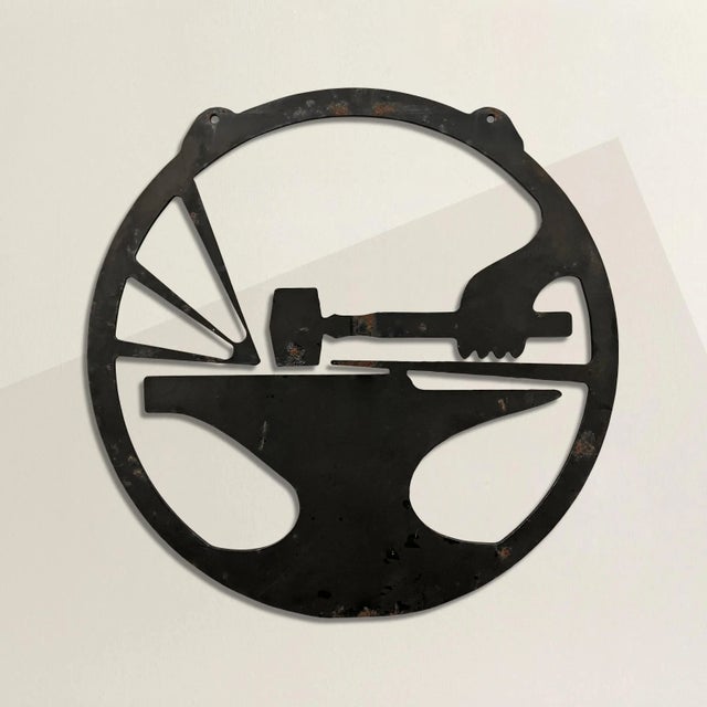 A bold and striking 20th century American blacksmith's sign with an arm swinging a hammer against a block of metal on an...