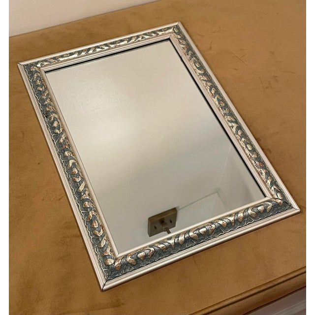 A raised ornate detail in silver finish with a light teal color underlay surrounds this wall mirror. The tone is soft...