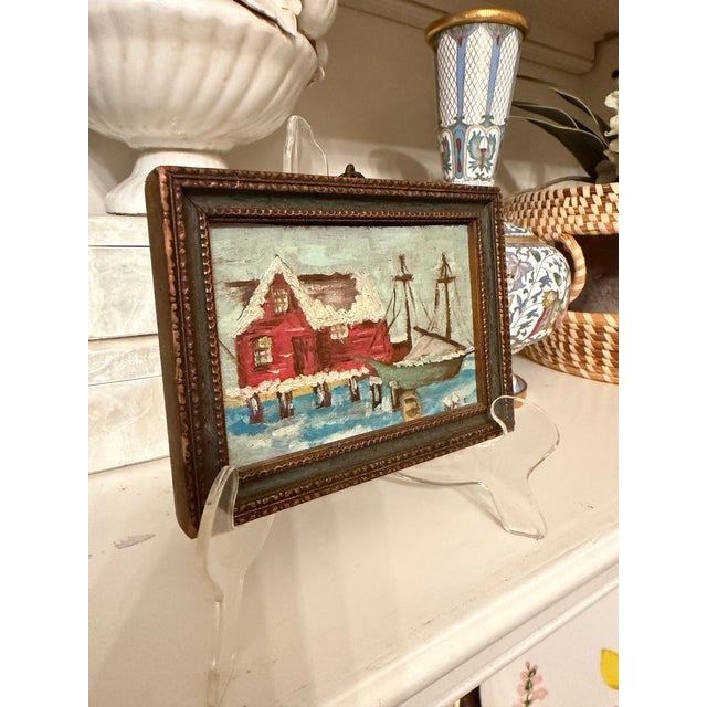 This charming small painting depicts a quaint coastal harbor scene, featuring a pair of red fishing cottages perched along...