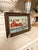This charming small painting depicts a quaint coastal harbor scene, featuring a pair of red fishing cottages perched along...