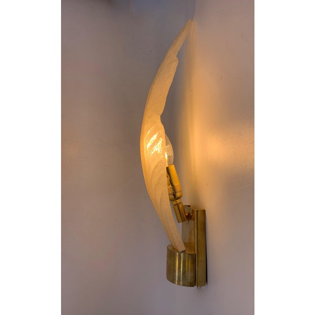The lamp is a decorative wall light with a sculptural design inspired by the shape of a stylized leaf. Crafted from clear...