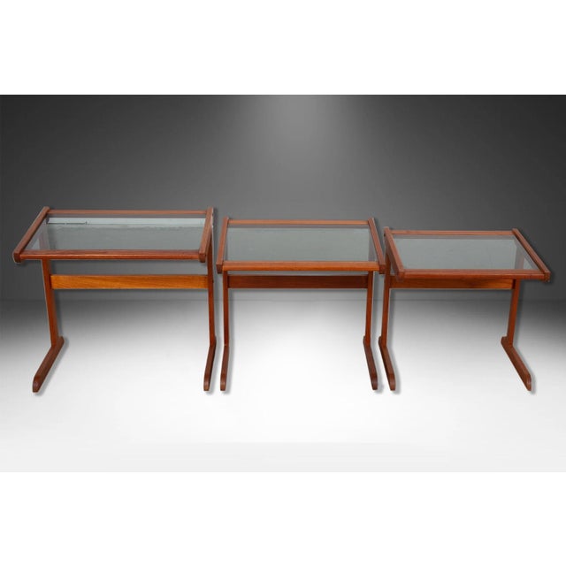 Set of three Mid Century Modern teak, rectangular smoked glass top nesting tables. The top rests on an attractive trestle...